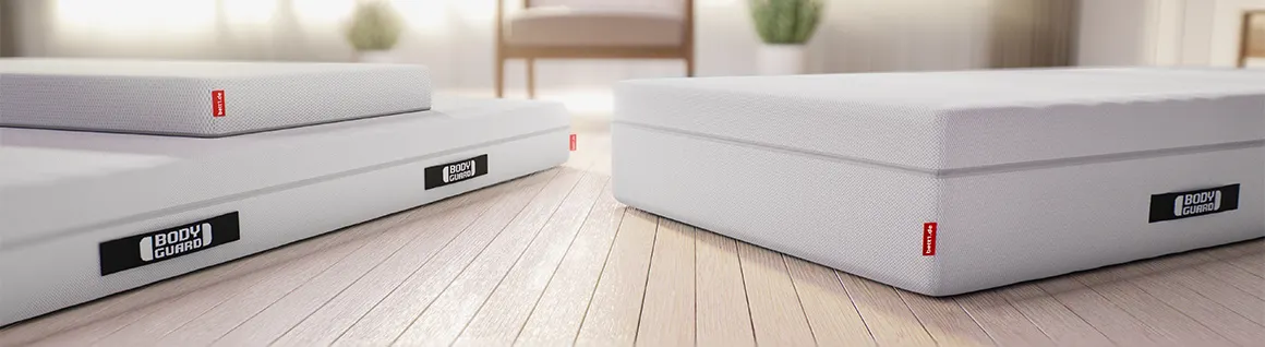 The bett1 mattresses are laid out on a wooden floor: on the left, the BODYGUARD Anti-Kartell-Matratze, on top of that the SuperBreeze Kids Mattress, and on the right, the BODYGUARD Box Spring Mattress.
