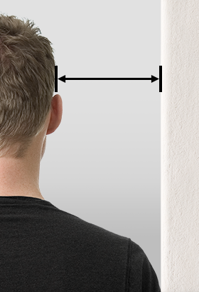 Foto: Eine Person steht mit der Schulter zur Wand, zwischen Wand und Ohr ist ein Pfeil eingezeichnet.