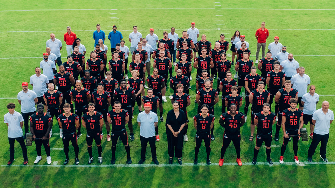 Photo: The Berlin Braves team in several rows on the pitch. The players in black and red jerseys, management and stuff around them in white tops.