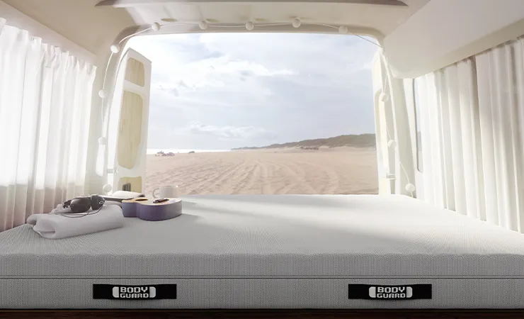 Interior view of a camper van containing a BODYGUARD mattress 140x190. Beach accessories, headphones, and a guitar are lying on the mattress. A beach can be seen through the open rear doors.