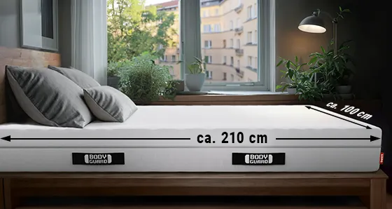 A BODYGUARD® mattress is lying in a bedroom. On the long side an arrow is labeled “approx. 210 cm”, on the wide side an arrow is labeled “approx. 100 cm”.