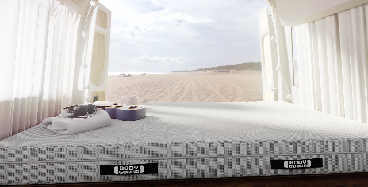 Interior view of a camper van with a BODYGUARD mattress measuring 150 × 200 cm. A towel, headphones, a coffee mug, and a guitar are lying on the mattress. A sandy beach is visible through the open rear doors.