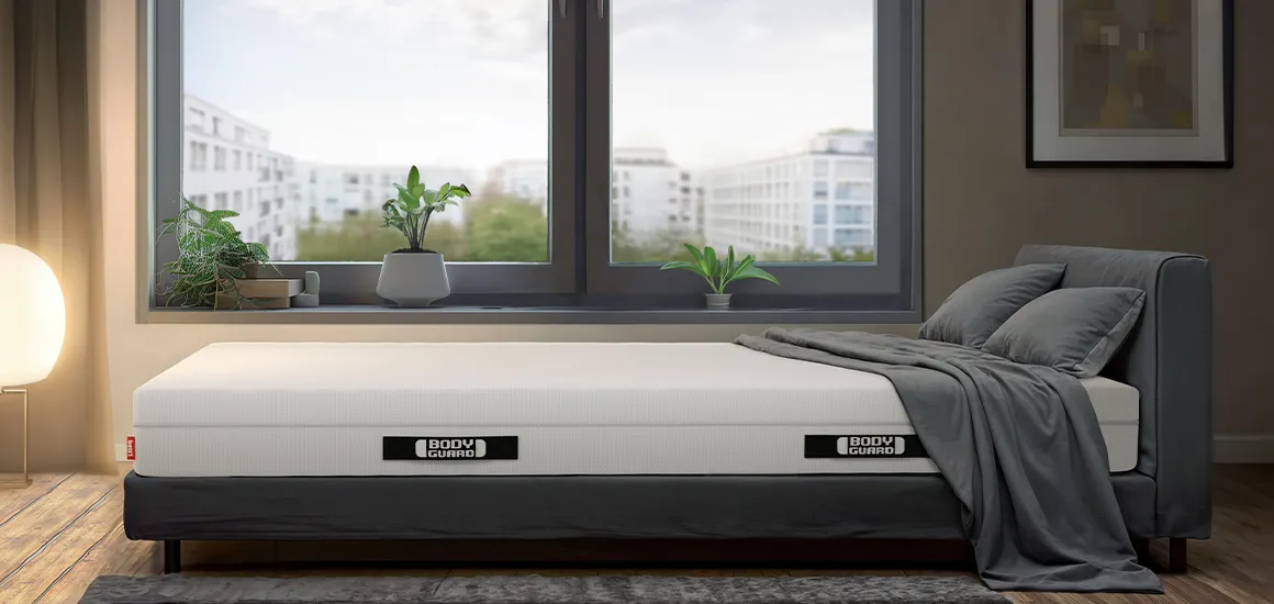 The BODYGUARD mattress on a bed in a bright bedroom. Behind it are large windows with a view of houses, and at the foot of the bed is a bright floor lamp.