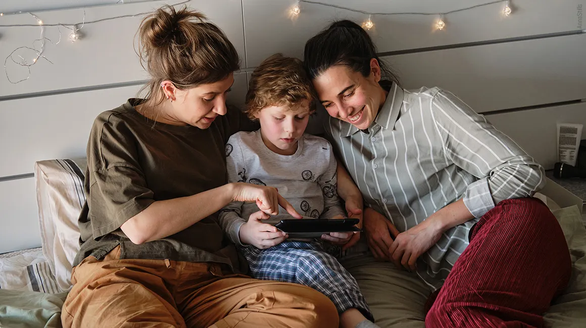 Photo: Two people and a child lying in bed, looking together at a tablet the child is holding.
