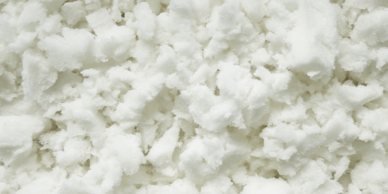Close-up of white memory foam flakes with a soft, fluffy texture. The irregularly shaped foam pieces appear light and airy and are densely packed together.