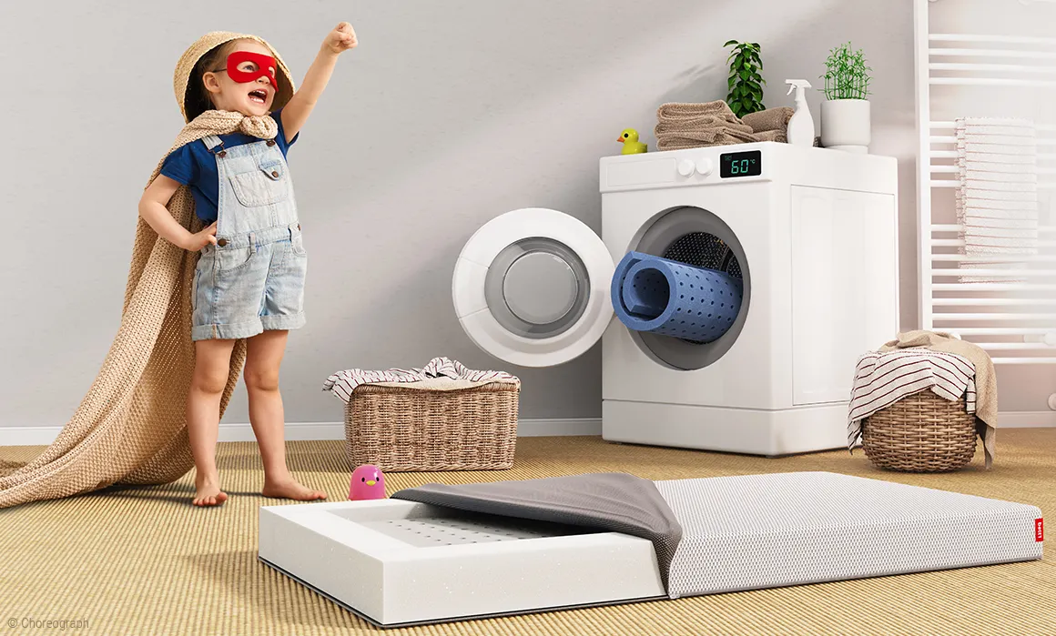 The BODYGUARD children's mattress lies on the floor next to a child in a superhero costume. The mattress cover is open, the removable lying surface has been removed and is rolled up in a washing machine behind the mattress.
