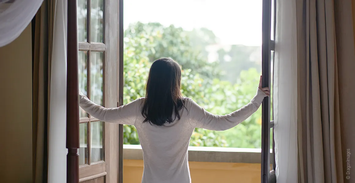 Photo: A person stands at a wide-open balcony door, arms on either side of the double doors, looking out at the greenery.