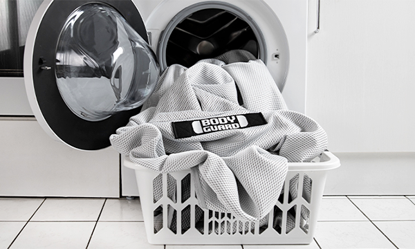 HyBreeze Cover with BODYGUARD logo placed in a laundry basket in front of a washing machine.