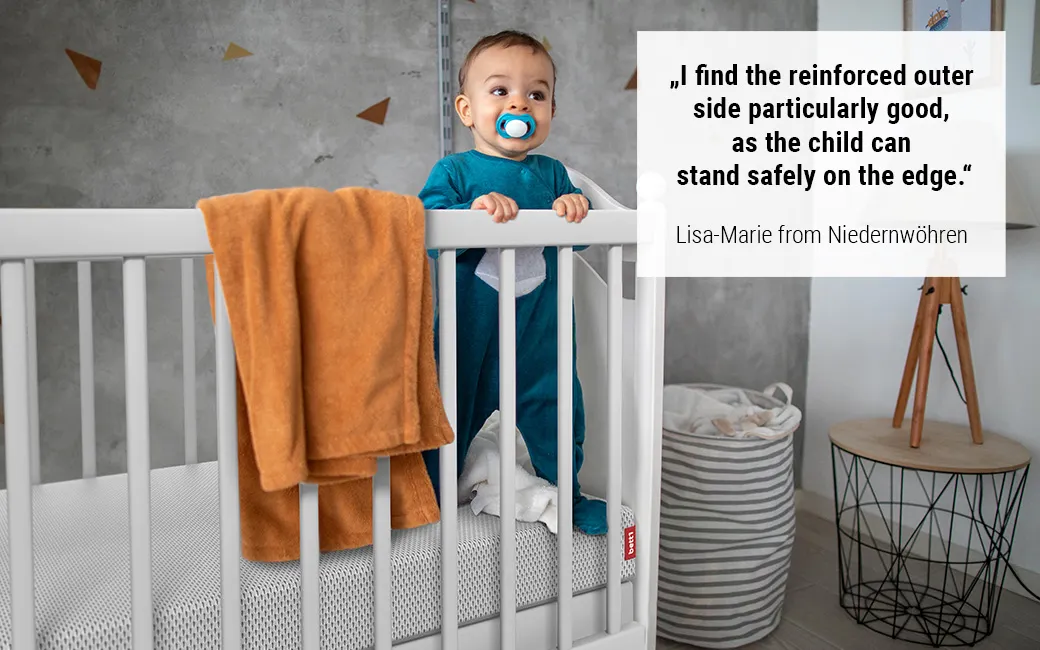 Baby stands in cot on the BODYGUARD® Kids Mattress. Text: "I particularly like the reinforced outer side, as the child can stand safely on the edge." Lisa-Marie from Niedernwöhren	