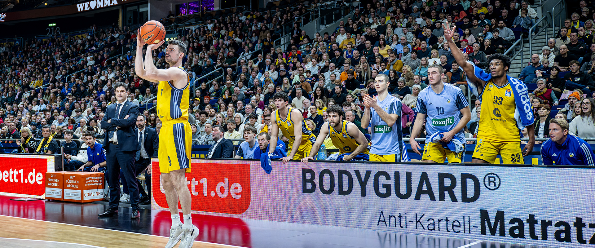 Photo: Game scene in a dark hall. Players in blue and yellow jerseys, one person throws the ball towards the basket.