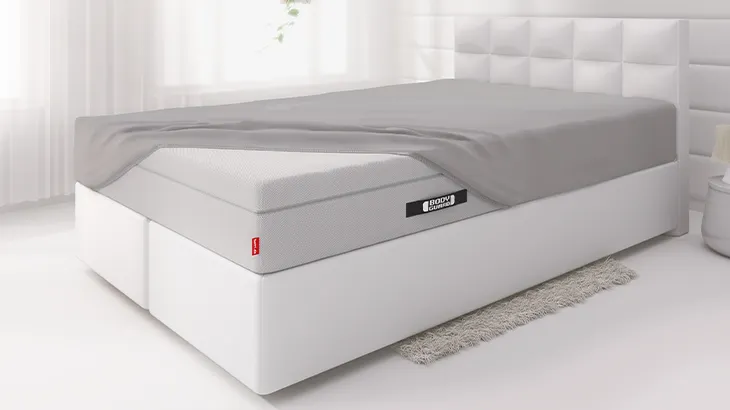 The BODYGUARD Box Spring Mattress lies on a light-coloured bed in a light-coloured bedroom. The mattress is covered with the BODYGUARD Fitted Sheet extra deep, except for one corner.