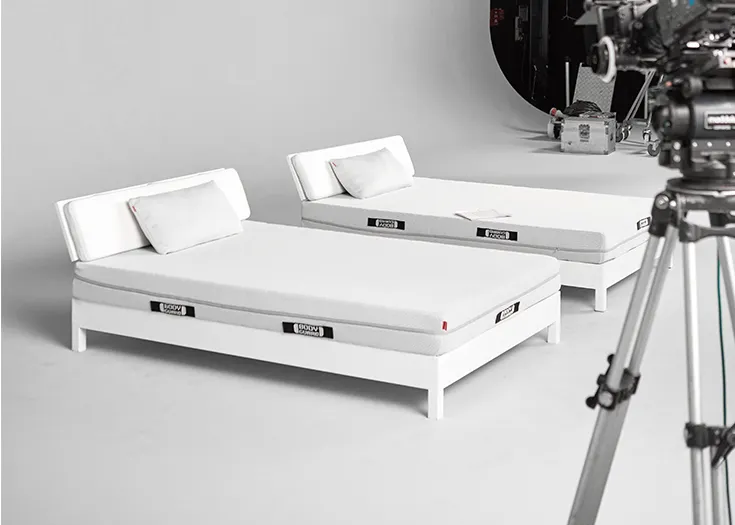 Photo: Two beds stand in front of a light-colored wall, each topped with a BODYGUARD mattress and a BODYGUARD visco pillow. In the foreground, a film camera on a tripod.