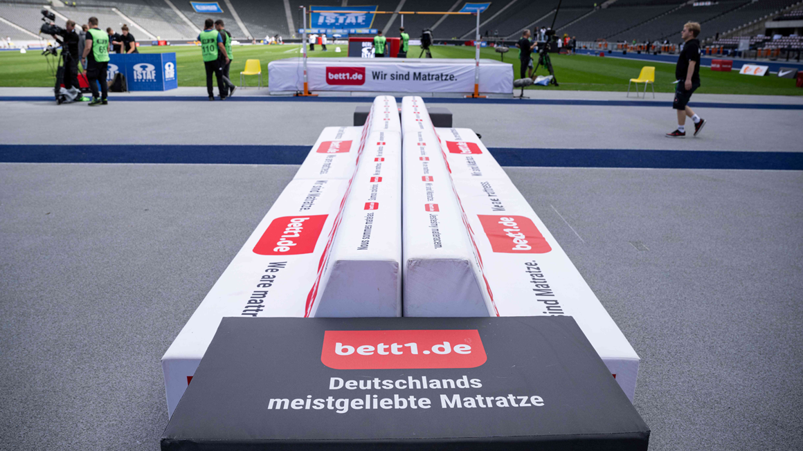 Photo from the edge of the pitch in a stadium, above a players' bench with bett1 logo on athletics stations in the middle of the pitch.