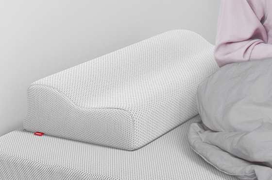 The BODYGUARD neck support pillow is lying on a BODYGUARD mattress, next to it is a person sitting halfway under a duvet.