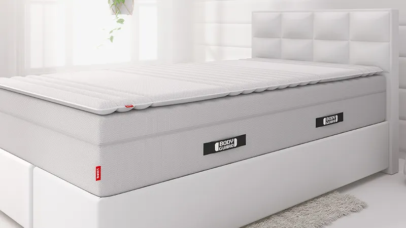 The extra-high BODYGUARD mattress lies on a white box spring bed, with the 140x200 cm topper on top.