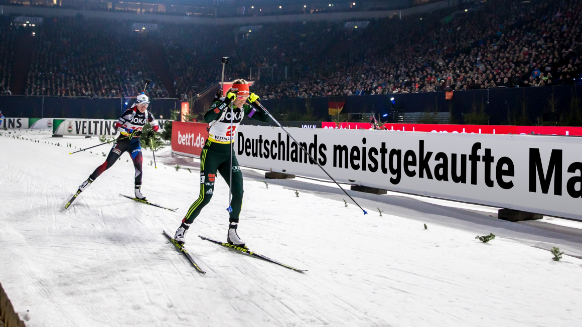 Photo: A few people on skis in a row on a running track in the stadium. The stadium banner reads in large letters: Germany's best-selling mattress.