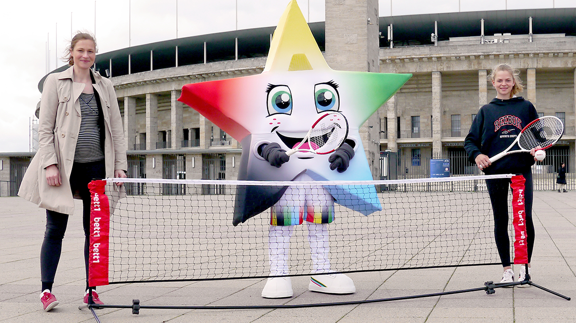 Photo: Two people and a mascot in a star costume stand with tennis rackets behind a small tennis net in front of the Olympic Stadium.