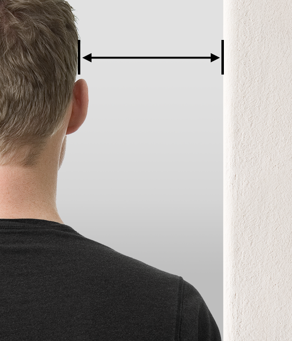 Foto: Eine Person steht mit der Schulter an einer Wand. Ein Pfeil zwischen Wand und Kopf deutet den Abstand an.