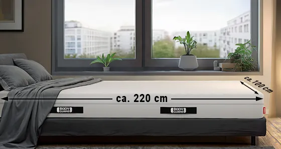 A BODYGUARD® mattress is lying in a bedroom. On the long side an arrow is labeled “approx. 220 cm”, on the wide side an arrow is labeled “approx. 100 cm”.
