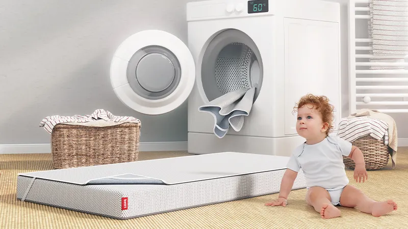 The BODYGUARD® Kids Mattress covered with a wetness protection pad lies in front of an open washing machine from which another wetness protection pad is hanging.