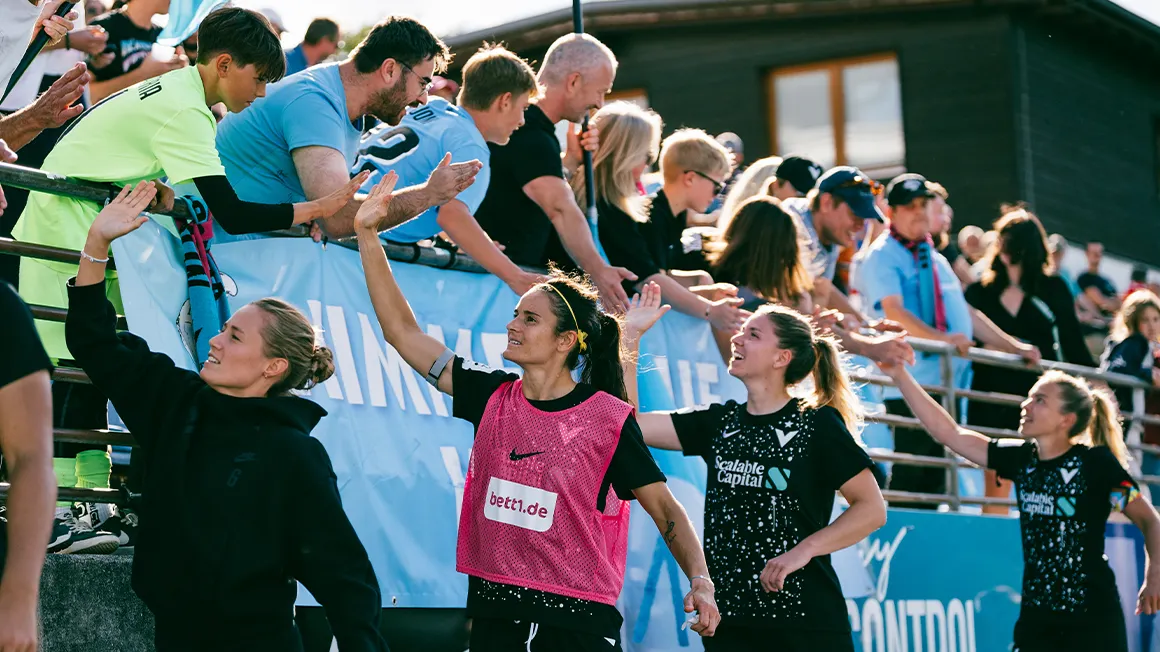 Foto: Spielerinnen des FC Victoria stehen unter der Tribüne und reichen dem Publikum die Hände.