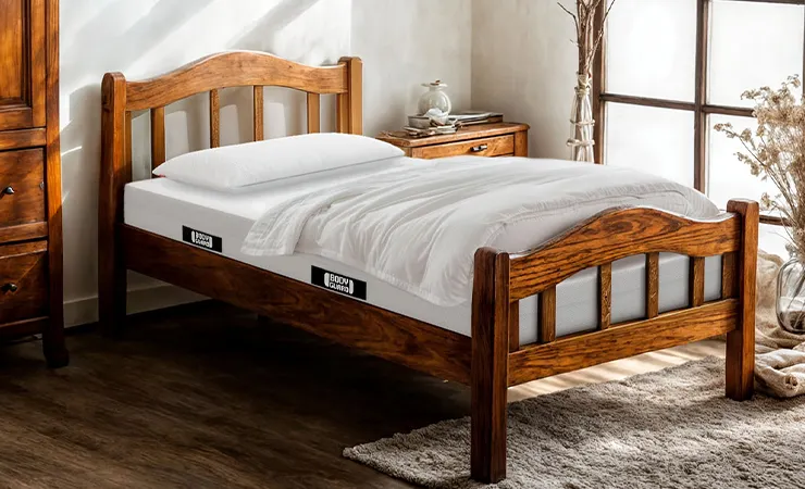 Ein helles, rustikal eingerichtetes Schlafzimmer mit viel Holz. In einem kurzen Bettgestell aus Holz liegt eine BODYGUARD Matratze 140x190 mit Kissen und Decke.
