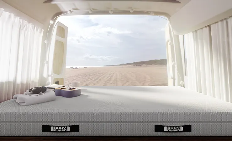 Innenansicht eines Camping-Vans, in dem eine BODYGUARD Matratze 140x190 liegt. Auf der Matratze liegen unter anderem Strandutensilien, Kopfhörer und eine Gitarre. Durch die geöffneten Hintertüren ist ein Strand zu sehen.