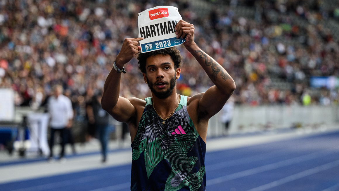 Photo: A runner on a tartan track holds a sign with his name and bett1 logo above his head with both arms.