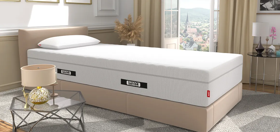 The BODYGUARD mattress on a bed in a bright bedroom. The BODYGUARD Visco pillow is on the mattress. In the background, floor-to-ceiling windows are open, offering a view of a city; in the foreground, there is a side table with a lamp.