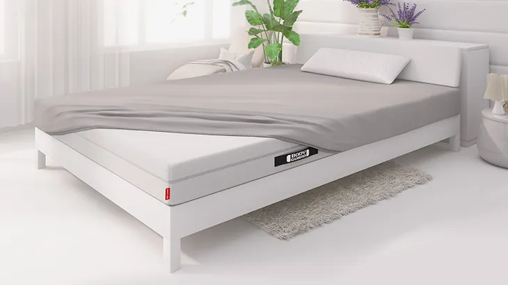 The BODYGUARD Anti-Kartell-Matratze lies in a light-coloured bedroom with a light-coloured bed. The mattress is covered with the BODYGUARD Fitted Sheet, leaving one corner uncovered.
