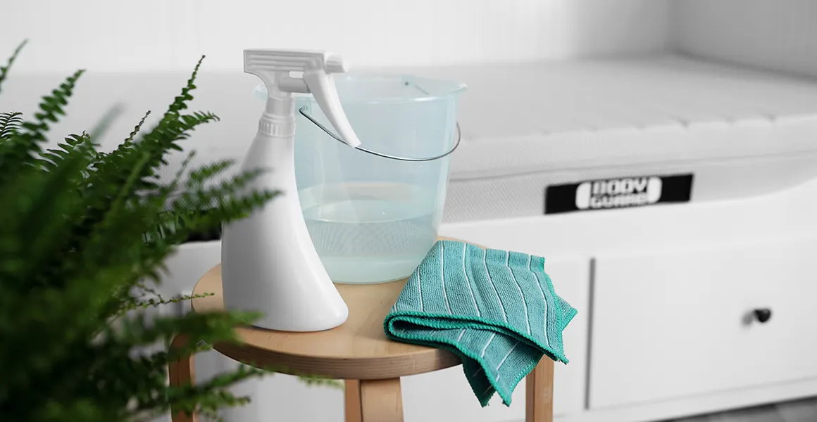 Photo: Cleaning supplies, a spray bottle, a microfiber cloth, and a bucket of water are standing on a stool, with a BODYGUARD mattress in the storage bed behind it.