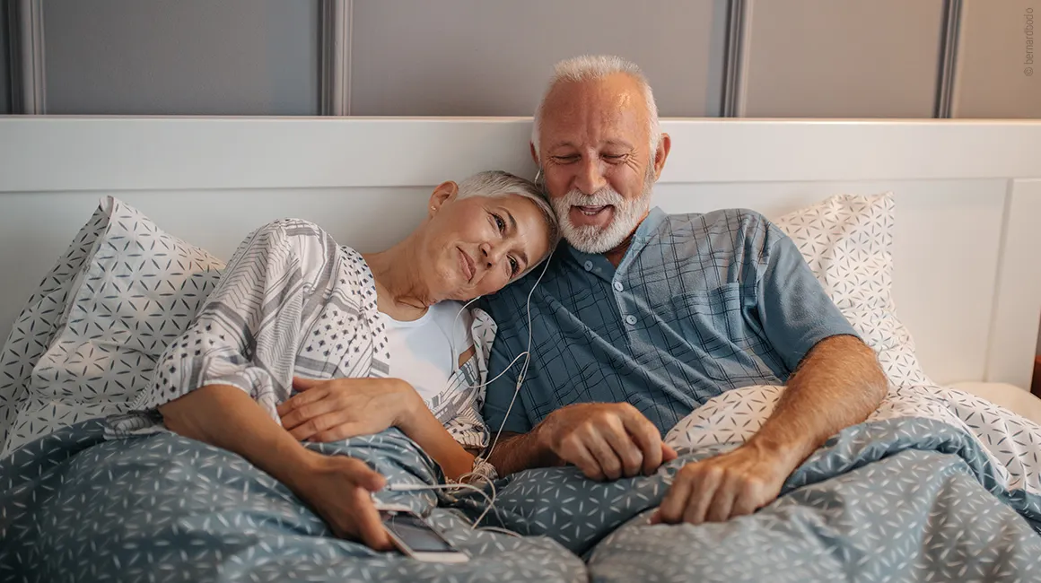 Photo: Two people sitting under a blanket in bed, listening to music through headphones. 