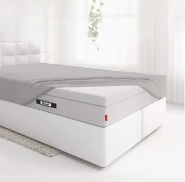 The BODYGUARD® Boxspring mattress is covered extra high except for one corner with the fitted sheet and lies on a boxspring bed in a bright bedroom.