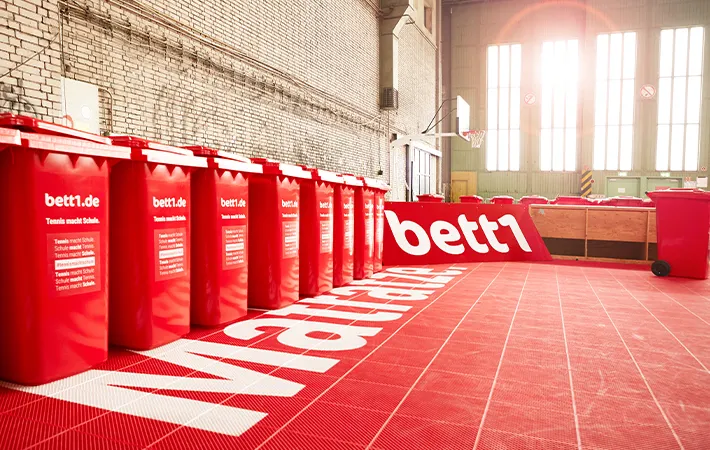 Foto: Eine Reihe von roten bett1-Tonnen auf einem roten Hallentennisplatz.