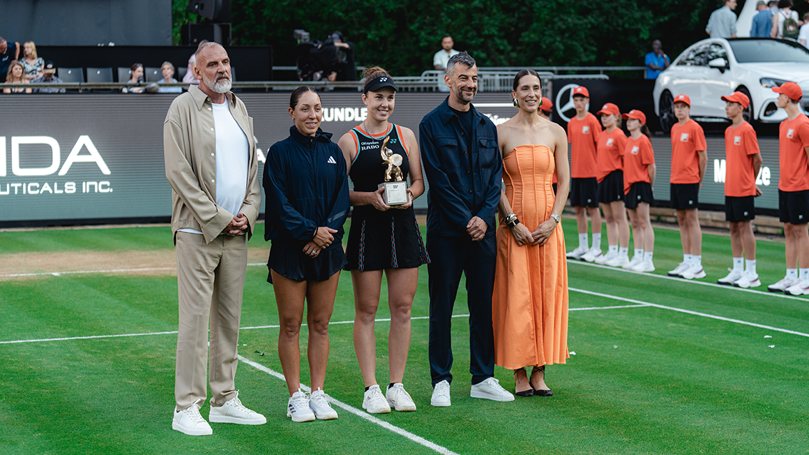 Five people are standing side by side on the tennis court. The one in the middle is holding a trophy.