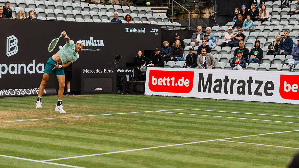 Auf einem Tennisplatz: Person schlägt einen Ball. Im Hintergrund sitzen Zuschauende auf der Tribüne, daneben sind bett1.de-Werbebanden zu sehen.