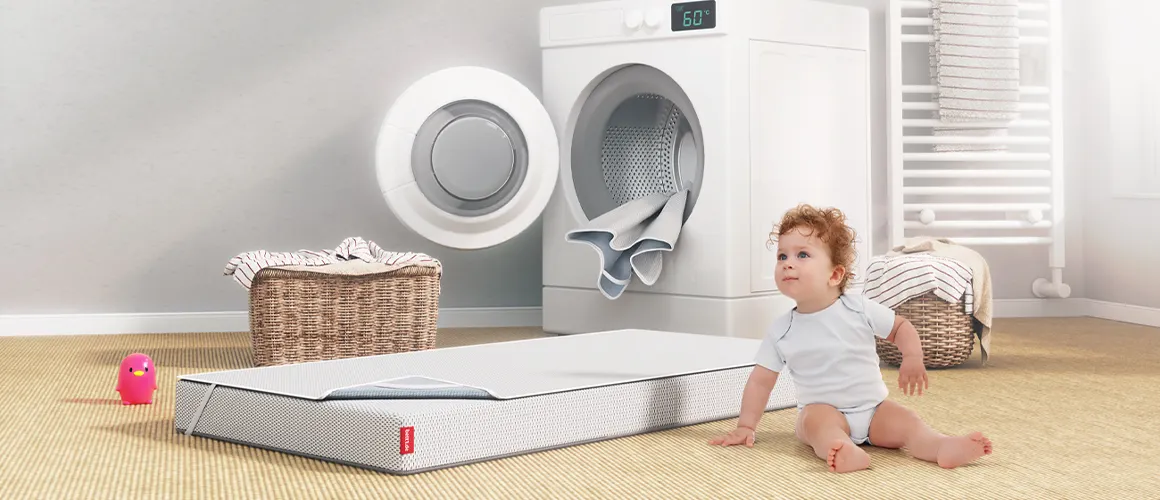 A toddler is sitting in a bathroom in front of a washing machine. Next to it, the SuperBreeze Kids Mattress lies on the floor. It is covered with the BODYGUARD Protector for Kids Mattresses.