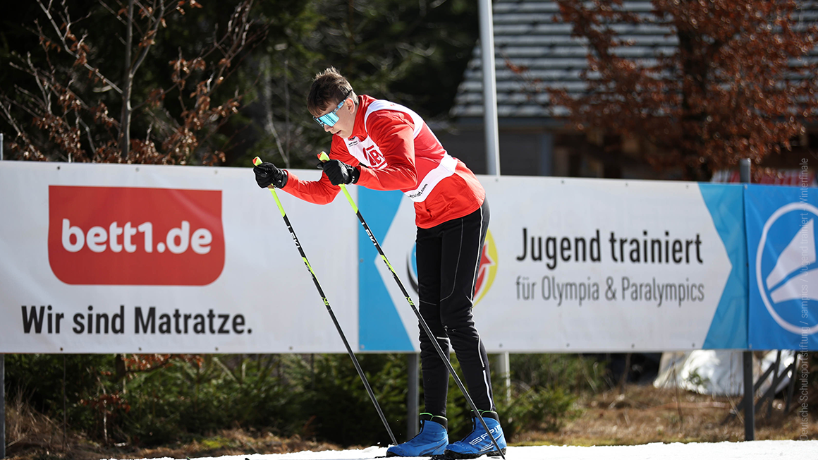 In front of an advertising hoarding, a person is standing on skis, leaning forwards, and planting their ski poles into the ground.
