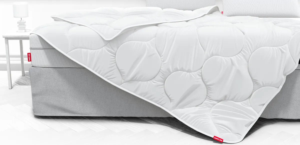 Ein hell eingerichtetes Schlafzimmer. Die BODYGUARD Ganzjahresdecke 155x220 cm liegt halb auf einem Boxspringbett. Es wirkt, als würde sie vom Bett gleiten. 