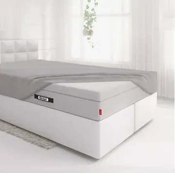 The BODYGUARD® Boxspring mattress is covered extra high except for one corner with the fitted sheet and lies on a boxspring bed in a bright bedroom.