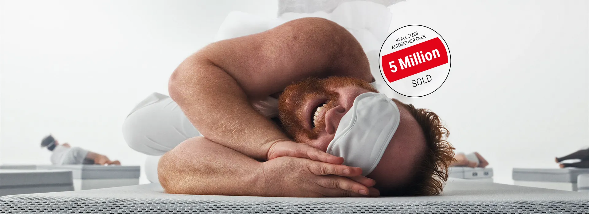 A man with a full red beard and a sleep mask lies on his side on a BODYGUARD mattress, smiling happily, with his palms pressed together beneath his face. Text: Over 5 million sold in all sizes.