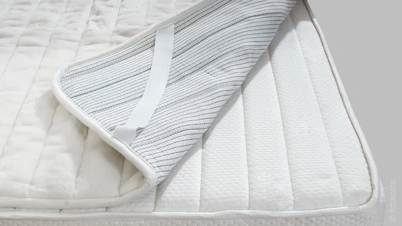Photo: A waterproof mattress topper lies on a mattress, one corner is folded back to reveal the fastening strap and waterproof coating.