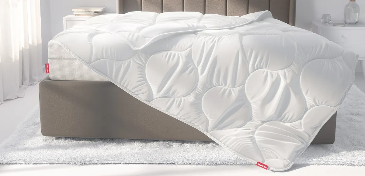 Ein hell eingerichtetes Schlafzimmer. Die BODYGUARD Ganzjahresdecke 135x200 cm liegt halb auf einem Boxspringbett. Es wirkt, als würde sie vom Bett gleiten.