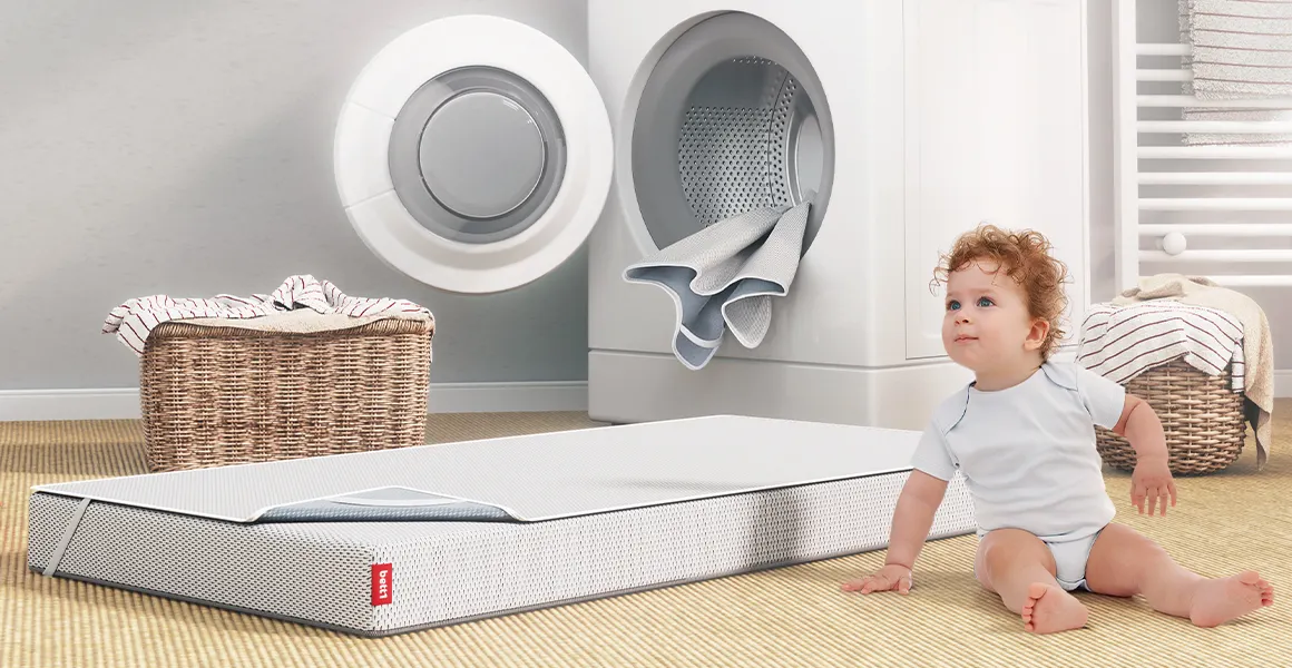 A children’s mattress with BODYGUARD Kids Mattress Protector is sitting in front of an open washing machine. Next to it there is sitting a toddler, looking on curiously.