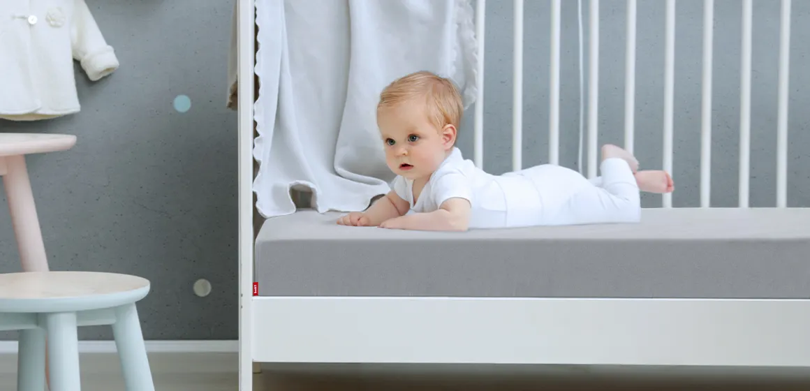 A baby is lying on their tummy in a cot. The cot is fitted with the SuperBreeze Kids Mattress and the BODYGUARD Fitted Sheet 70x140 for children.