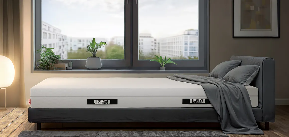 The BODYGUARD mattress on a bed in a bright bedroom. Behind it are large windows with a view of houses, and at the foot of the bed is a bright floor lamp.