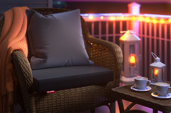 The RAINGUARD Seat Cushion is sitting on a garden armchair outside in the evening light. In the background is a railing with fairy lights. In the foreground is a side table.