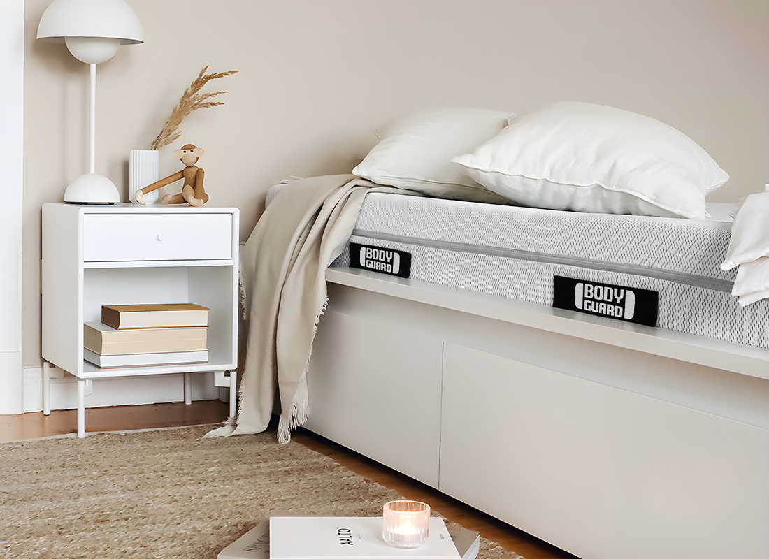 A bright room. A BODYGUARD mattress lies in a bed, next to it a bedside table with a wooden monkey