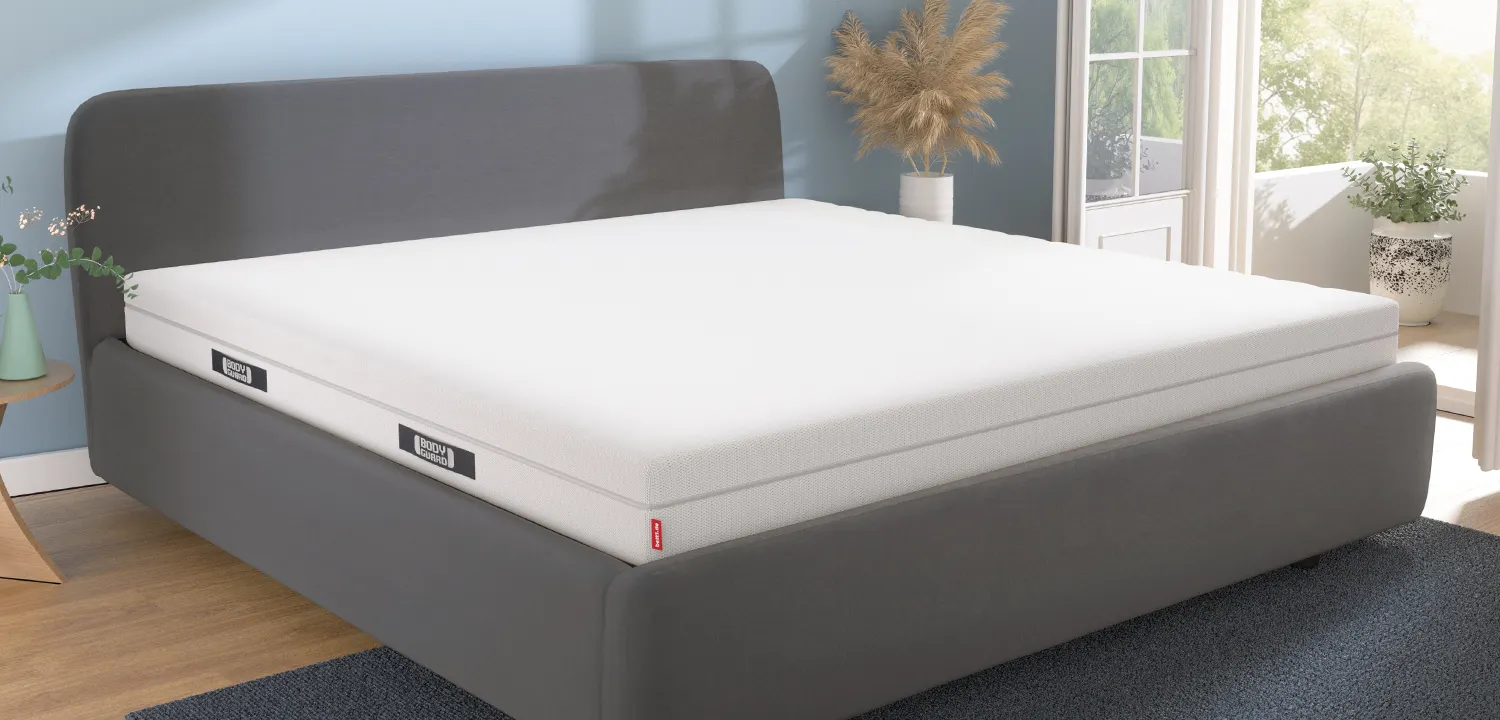 Bett mit BODYGUARD Matratze 180x220 cm in einem hellen, modern eingerichteten Schlafzimmer. Durch die geöffnete Balkontür scheint die Sonne herein