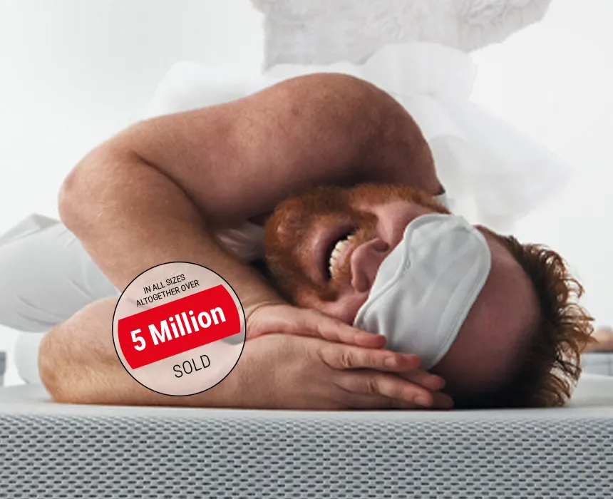 A man with a full red beard and a sleep mask lies on his side on a BODYGUARD mattress, smiling happily, with his palms pressed together beneath his face. Text: Over 5 million sold in all sizes.